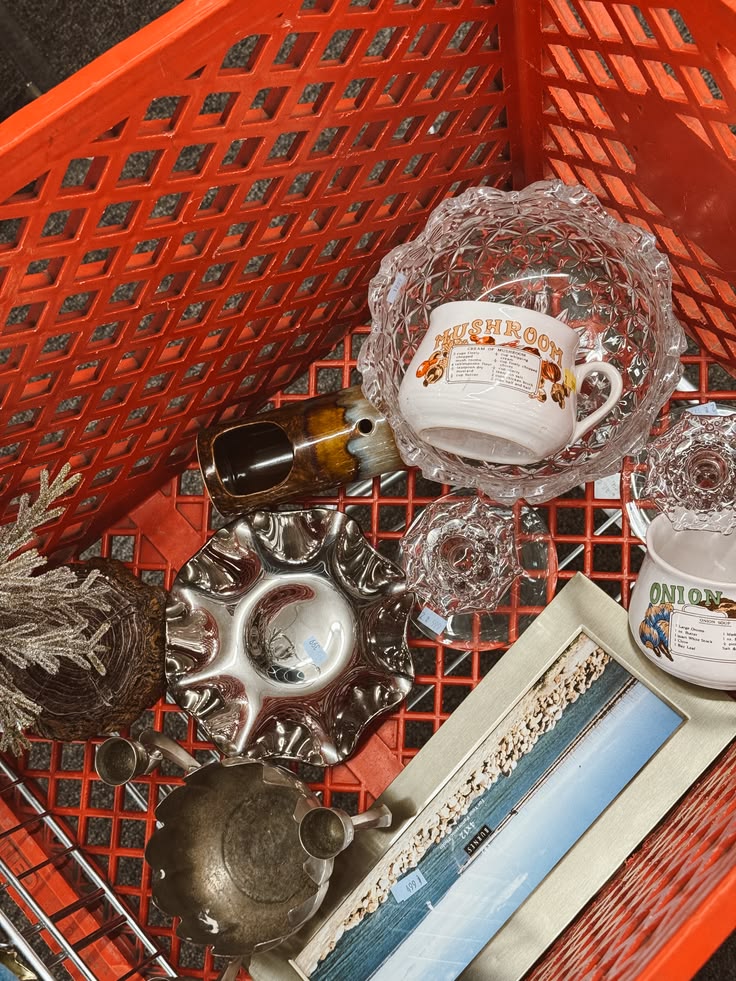 A red thrift basket filled with vintage glassware, mugs, and finds